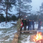 Grupa ludzi stoi przy ognisku w zaśnieżonym, zalesionym terenie. W pobliżu zaparkowane są rowery. Niektórzy ludzie mają na sobie zimowe ubrania, a za nimi na drążkach zamontowany jest rower. Scenę otaczają wysokie drzewa.